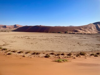 Vast dry clay pan surrounded by towering red sand dunes under a clear blue sky in Namibia's Namib...
