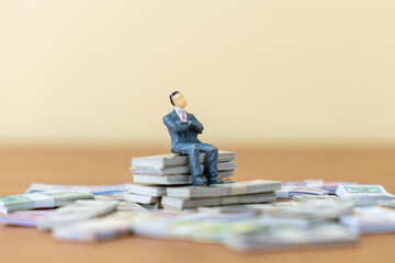 A miniature businessman sits on top of a large stack of US dollar bills, surrounded by other...