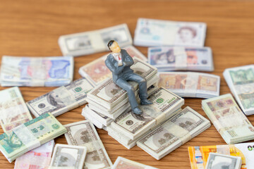 A miniature businessman sits on top of a large stack of US dollar bills, surrounded by other international currencies. Wealth, financial success, global investments, international business concept.