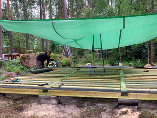 Crafting a rustic forest sanctuary, a focused Middle-Eastern man assembles a deck under a verdant shade tarp, celebrating International Building Safety Week