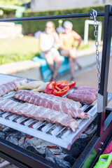 Grilling ribs, chicken and sausages with vegetables on barbecue with friends in background
