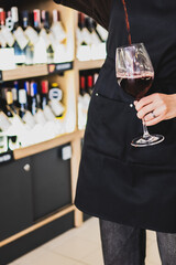 woman in a black apron pouring red wine into a glass in a wine shop, with shelves of bottles in the...