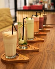 Aesthetic Drinks Row at the Cafe
A row of six drinks in various colors are served on a special wooden tray, arranged diagonally on a wooden table. Starting from milkshakes, mocktails, smoothies, and i