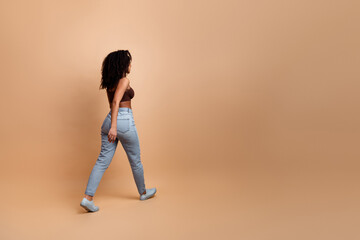 Young woman in casual outfit against beige background walking gracefully highlighting trendy...