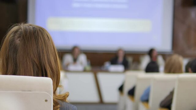 Woman sits in a classroom with a projector screen behind her. The projector screen shows a presentation with a woman in the foreground. The classroom is full of people