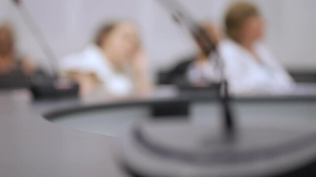 Blurry image of three people sitting at a table with a microphone in front of them. The people are wearing white shirts and the microphone is black. Concept of formality and professionalism