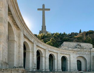 Stone archways and cross monument with hills and trees under a clear blue sky, Basilica of the Holy Cross of the Valley of Cuelgamuros, Madrid, Spain