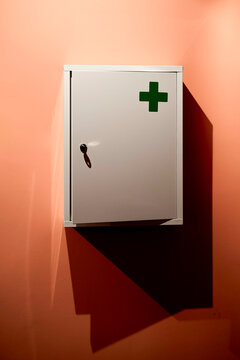 White first aid cabinet with green cross on peach wall casting a shadow. Ghent, Belgium