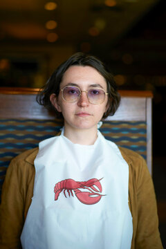 Young woman wearing glasses and white bib with red lobster print, seated indoors with blurry background.   Palm Springs, USA
