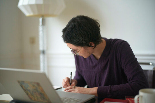 Woman focused on writing at a desk with a laptop and lamp in a calm setting.