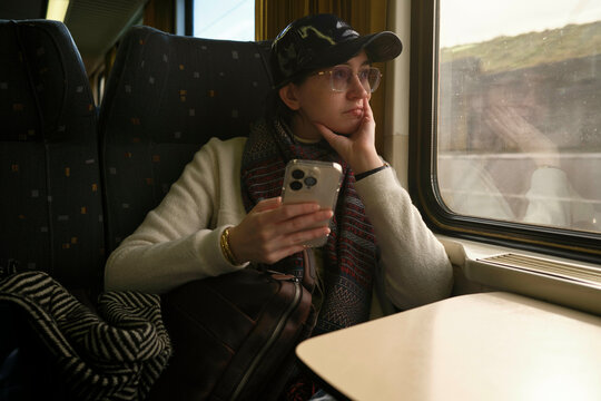 Woman in a train seat gazing out the window while holding a smartphone.