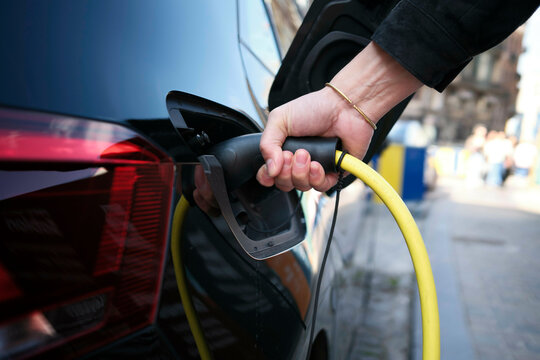 Hand plugging a charger into an electric car on a street in daylight.