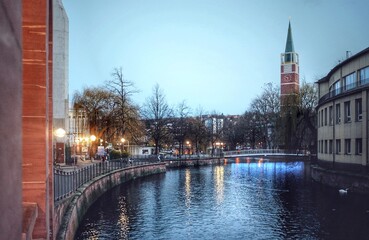 canal with glowing evening reflections, buildings and trees on the banks, and a clock tower in the...