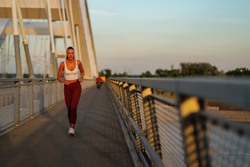 Athletic woman running on a bridge during sunset, with a focus on fitness and health. The warm glow...