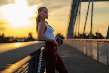 Young athletic woman enjoying a breathtaking sunset view from a sleek modern bridge, holding her...