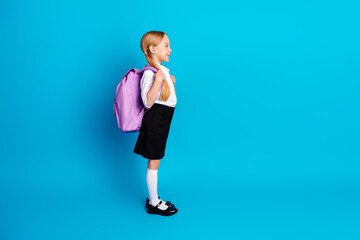 Adorable young schoolgirl with backpack standing on vibrant blue background, ready for school with...