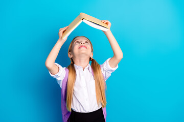 Happy schoolgirl holding a book on her head against a blue background, expressing excitement and...