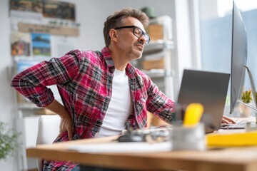 Obraz premium Stressed businessman suffering from back pain while working at home office desk, side view.