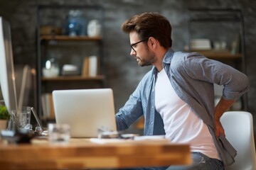 Fototapeta premium man with back pain sitting at a desk in a home office, side view. High-quality photo of a business person suffering from upper body discomfort due to prolonged work on a computer screen at a table.