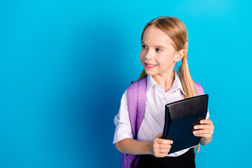Young schoolgirl with a purple backpack holding a tablet device against a blue background, smiling...