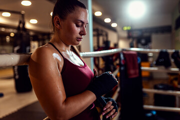 Female boxer leaning against the ropes of a boxing ring wrapping hands with boxing strap.