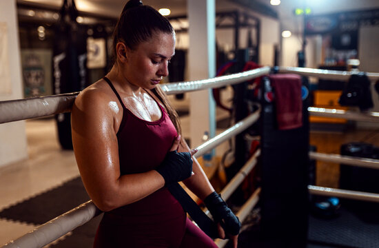 Female boxer leaning against the ropes of a boxing ring wrapping hands with boxing strap. - Powered by Adobe