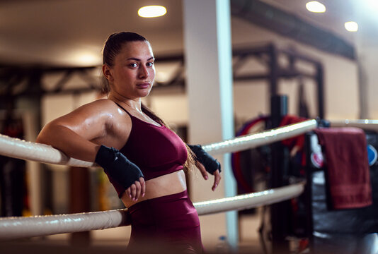 Female boxer leaning against the ropes of a boxing ring resting after training looking at camera.