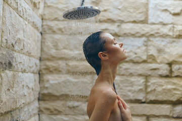 woman showering under modern rain showerhead with stone wall background, calm expression, natural...