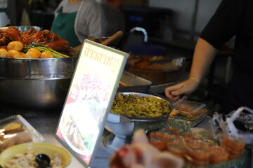 Street food vendor preparing stewed pork leg with vegetables and rice, with a glowing menu sign and condiments in the foreground.