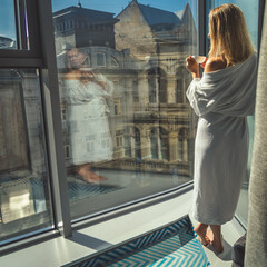 Woman in elegant robe drinking coffee in hotel room and standing near window