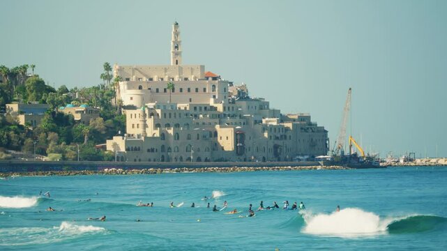 View on old historic port city of Jaffa in Tel Aviv with surfers in sea water. Summer day outdoors in Israel famous city by the Mediterranean Sea. People enjoy surfing and swimming by picturesque view