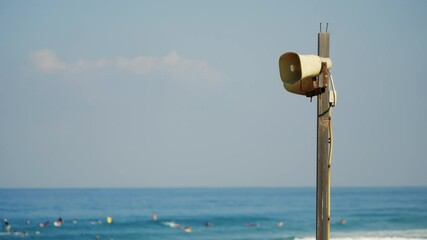 Outdoor loudspeaker or megaphone for public announcements siren or alarm. Installed on wooden pole stand is loudspeaker by ocean or beach with swimmers and bathers in water in background on summer day - Powered by Adobe