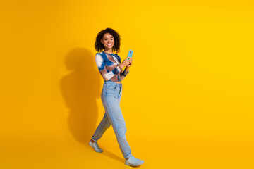 Young woman with curly hair holding a smartphone, smiling confidently in casual fashion against a...