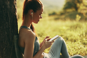 Young woman enjoying music outdoors in athletic wear, sitting under a tree, smartphone in hand, natural setting, serene atmosphere, healthy lifestyle