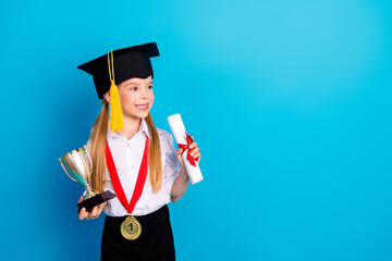 Adorable young girl in graduation cap holding a trophy and diploma on a blue background,...