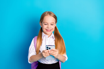 Cheerful schoolgirl in uniform holding a smartphone against a blue background, showcasing modern...