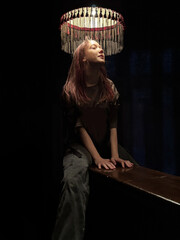 Mystical portrait of teenage girl with long red hair under glowing vintage lampshade in dark room