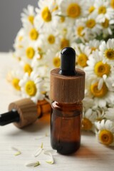 Bottle of essential oil and chamomile flowers on light wooden table, closeup