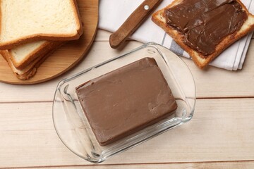 Block of tasty chocolate butter in keeper and slices of bread on light wooden table, flat lay