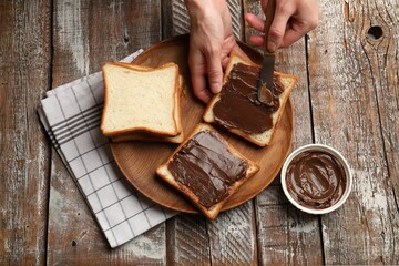 Woman spreading tasty chocolate butter onto bread at wooden table, top view