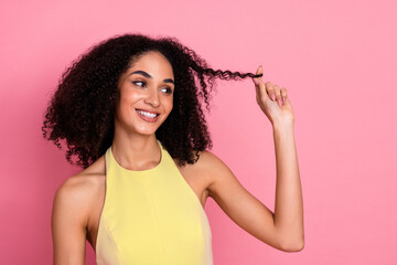 Smiling young woman with curly hair playing with her locks in a yellow dress against a vibrant pink...