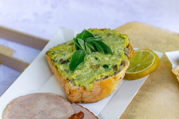 Herb spread toast garnished with fresh leaves, served with a slice of lemon and cold cuts on a white plate – a simple rustic-style snack.