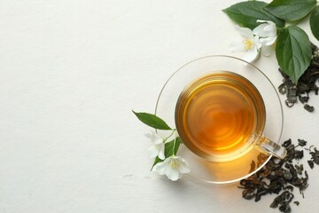 Aromatic jasmine tea in glass cup, flowers and leaves on white table, flat lay. Space for text