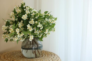 Beautiful jasmine flowers in vase on wicker table near window indoors, space for text