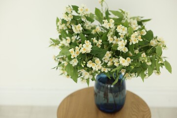 Beautiful jasmine flowers in vase on wooden table near white wall indoors, closeup. Space for text