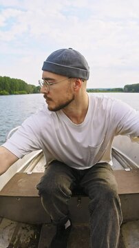 Attractive man, fisherman with beard and dockworker's hat, is rowing boat with two hands in middle of the river during the summer.