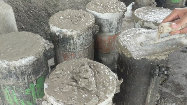 Worker leveling fresh concrete in cylindrical molds for construction material testing in a laboratory setting.
