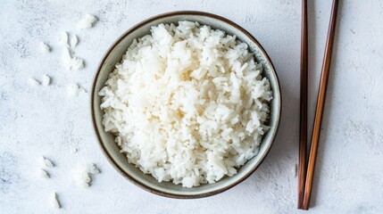A bowl of plain cooked white rice, accompanied by chopsticks, rests on a mottled, light-grey surface.  A few stray grains are scattered nearby