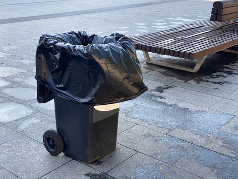 Trash can with black bag stands near wooden bench. Cleanliness of city. Waste sorting. 