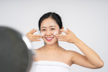 Smiling cute Asian girl looking at the mirror, her hand is applying moisturizer lotion on her face